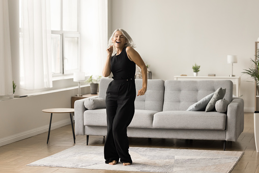 Joyful active senior lady excited with single dance at home enjoying motion, activity, music, singing song, having fun in cozy comfortable living room interior. Full length wide shot