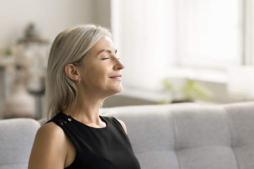 Happy relaxed mature woman enjoying fresh air, smells, relaxation, leisure at home, taking deep breath with closed eyes, smiling, meditating, reloading mind, enjoying stress relief technique