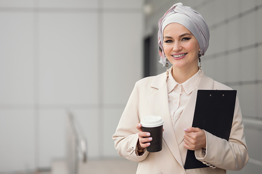Muslim woman smiles standing near office building. Woman wearing headscarf glad to meet friend. Businesswoman in formal clothes holds coffee cup and clipboard