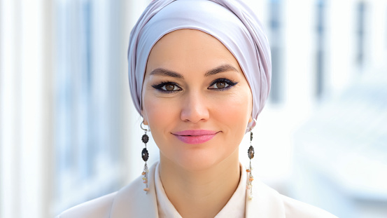 Muslim businesswoman wearing silver headscarf raises head and looks smilingly against grey city building. Woman in suit enjoys life of rich entrepreneur