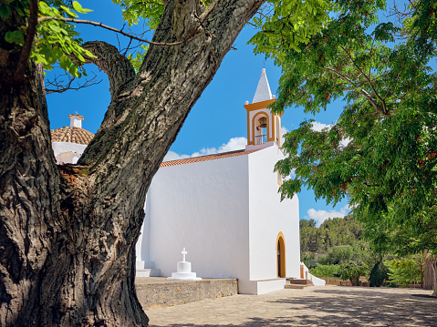 Wide-angle view of the church and town square of Sant Joan de Labritja (San Juan Bautista), located in the northern inland of Ibiza, built in the typical white lime style of the island and considered one the best examples of the island's traditional architecture. The dazzling bright light of a Mediterranean summer noon, blue sky, picturesque clouds, lush trees. High level of detail, natural rendition, realistic feel. Developed from RAW.