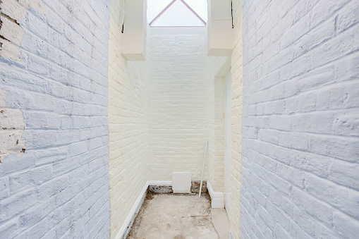Narrow hallway with white brick walls, skylight illumination, staircase with triangle-shaped window, eclectic designs.