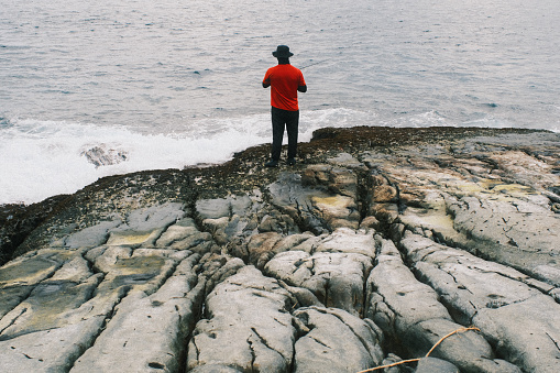 Balancing on the rocky shoreline, a man enjoys the solitary pursuit of fishing, his silhouette a testament to the tranquility and focus of this outdoor pastime.