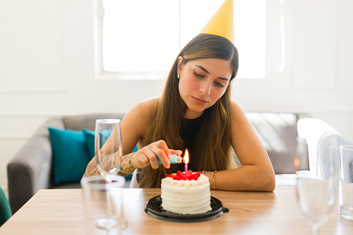 Upset sad beautiful woman lighting her candle on her birthday cake looking depressed celebrating her birthday alone with a party hat