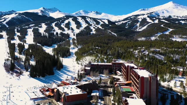 Chairlift at the Bottom of Ski Slope in Breckenridge, Colorado with People Skiing and Snowboarding next to Hotels and Resort. Aerial Drone Video