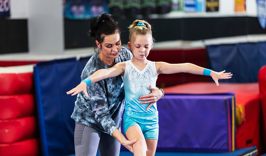 A 9 year old girl learning gymnastics. Her coach, a mature woman in her 40s, is helping her on the balance beam.