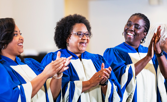 A group of three women having fun singing in a church choir wearing blue and white robes. The focus is on the woman in the middle. She is a senior, in her 60s.