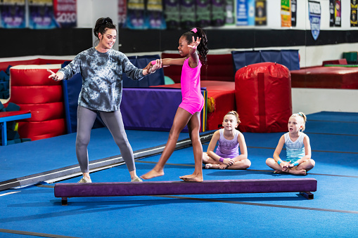 A 10 year old African-American girl learning gymnastics. Her coach, a mature woman in her 40s, is helping her on the balance beam. Two girls wait their turn in the background.