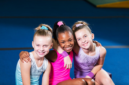 A multiracial group of three young gymnasts sitting side by side, smiling at the camera with their arms around each other's shoulders. The African-American girl is 10 years old, and her friends are sisters, 9 and 11.