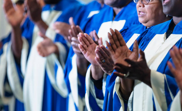 Cropped view of black men and women singing in choir Cropped view of a group of black and African-American men and women, mostly seniors, singing in a church choir, wearing blue and white robes. The focus the hands of the senior woman whose face is visible. black gospel church stock pictures, royalty-free photos & images