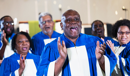 A group of senior black and African-American men and women singing in a church choir, wearing blue and white robes. The focus is on the man in the foreground. He is in his 70s.