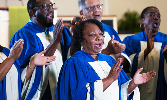 A group of senior black and African-American men and women singing in a church choir, wearing blue and white robes. The focus is on the woman in the foreground. She is in her 60s.