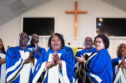 A group of mature and senior black and African-American men and women singing in a church choir, wearing blue and white robes. The focus is on the woman in the foreground who is in her 60s.