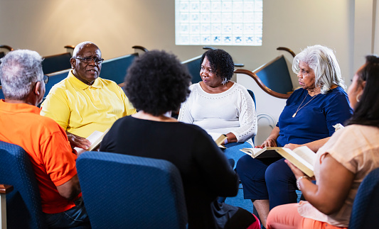A group of six men and women, mostly seniors, participating in bible study at church. sitting in a circle having a discussion. One of the men is speaking, leading the discussion. He is in his 70s.