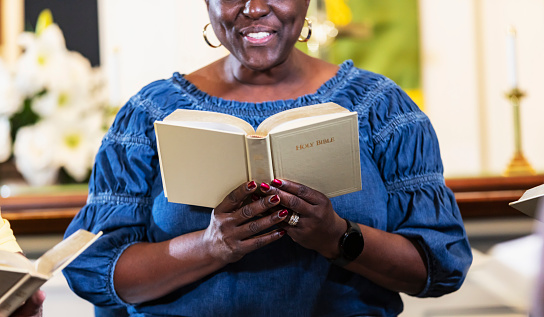 Cropped view of a mature black woman, in her 50s, sitting in a church, attending a bible study meeting. She reading the bible which she is holding open in front of her.