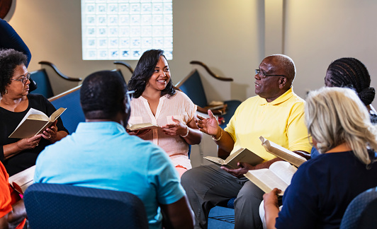 A group of men and women, mostly seniors, participating in bible study at church. sitting in a circle having a discussion. A senior man in his 70s is speaking with a serious expression. The woman next to him who is smiling is the youngest of the group, in her 30s.