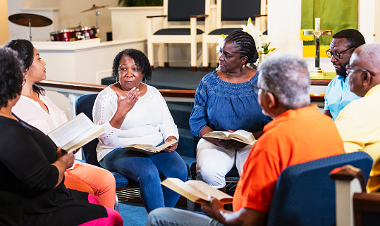 A group of men and women, mostly seniors, participating in bible study at church. sitting in a circle having a discussion. A senior woman is speaking with a serious expression and the others are looking toward her.
