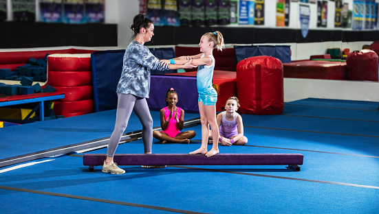 A 9 year old girl learning gymnastics. Her coach, a mature woman in her 40s, is helping her on the balance beam. Two girls wait their turn in the background.