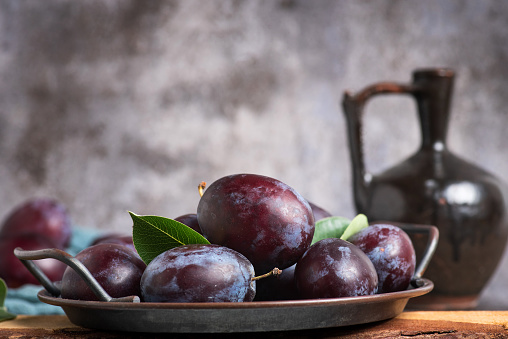 Ripe purple plums with leaves in a metal bowl on a dark wooden background