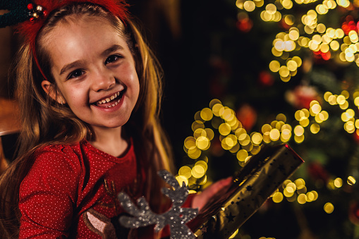 Portrait of beautiful little girl standing by the glistening Christmas tree, smiling at camera with joy.