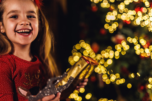 Portrait of beautiful little girl standing by the glistening Christmas tree, smiling at camera with joy.