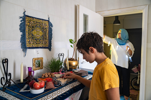 Photo of mother and 10 years old son decorating home for Nowruz Holiday. Nowruz taple is seen on foreground with mirror, candle, book, grains, wheatgrass  etc. Shot indoor under daylight.