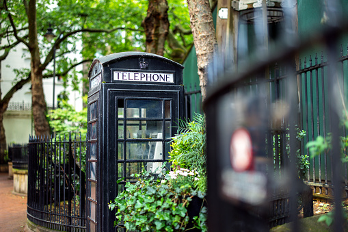 A classic black telephone booth in London located in a cute park. The telephone booth is surrounded by beautiful green trees and bushes.