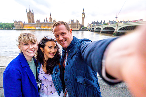 A group of three caucasian tourists visiting London together for the first time. They are taking a selfie in front of the Palace of Westminster and looking at the camera. They are standing by the river Thames. They are enjoying their time together exploring the city.