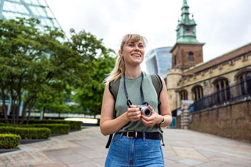 A young adult caucasian female tourist holding a camera in her hands as she is exploring the city of London. She is walking on a pedestrian street near a green park. The woman looks happy and is smiling as she is looking around and admiring the city. She is dressed in casual clothes and is wearing a ponytail.