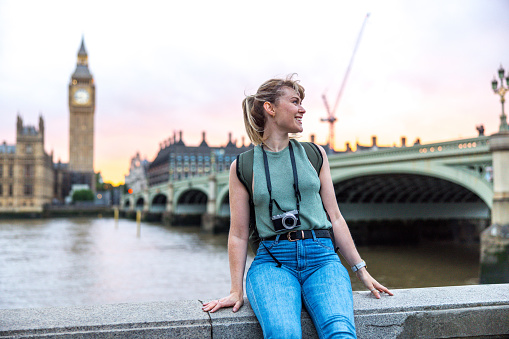 Happy young adult caucasian female exchange student spending her first day in London. She is posing for a photo by the river Thames with the view of Big Ben behind it. She is sitting on the concreate fence and looking away while smiling. Her camera is strapped around her neck. The sky is beautiful since the sun is setting.