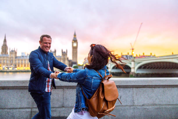 Cute Caucasian Adult Couple Holding Hands And Spinning In Front of The Elizabeth Tower As The Sun Is Setting A happily in love caucasian adult couple having fun together while exploring London during a beautiful sunset. They are holding hands and spinning in circles while looking at each other. They are located by the river Thames. The couple looks happy as they are being playful in front of the Elizabeth Tower. big ben stock pictures, royalty-free photos & images