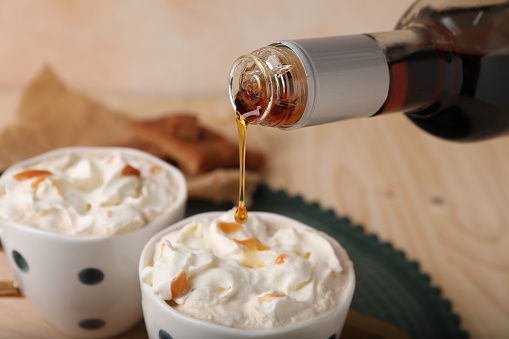 Pouring delicious caramel syrup into cup with coffee and whipped cream at wooden table, closeup