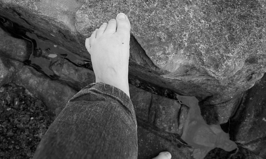 barefoot walking on dramatic rocks, pinky toe bandaged and curled under, top view of left foot, looking down, woman wearing blue jeans, we see the bottom of the jeans legs, black and white photo, small pale white woman's foot