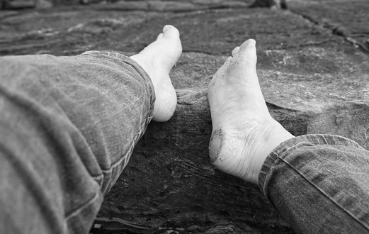 bare feet, dirty soles from walking barefoot, feet turned inward, resting on rugged rocks at the beach, looking down, close in, woman wearing blue jeans, we see denimum clad legs outstreched in black and white, small pale white woman's foot