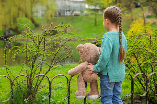 Little girl with teddy bear outdoors, back view. Space for text