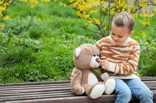 Little girl with teddy bear on wooden bench outdoors. Space for text
