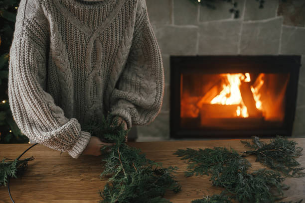 istock Hands in cozy sweater making Christmas rustic wreath with fir branches on wooden table against cozy fireplace, close up. Top view. Winter holiday preparations, atmospheric time 1709202354