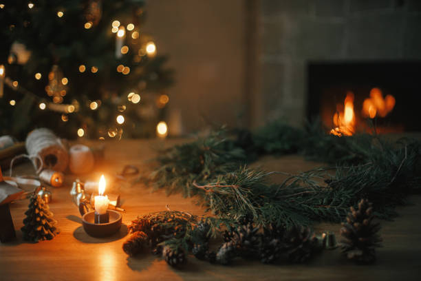 haciendo una corona rústica navideña. ramas de cedro, piñas, velas, cordeles, campanas sobre mesa de madera contra árbol y chimenea en sala de noche. preparativos para las vacaciones de invierno, tiempo atmosférico - fireplace christmas candle holiday fotografías e imágenes de stock