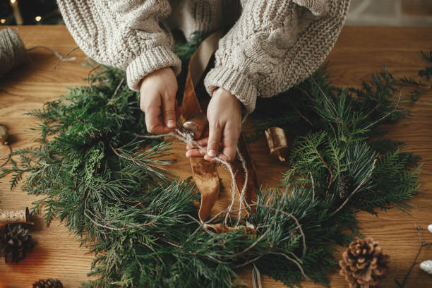 Making Christmas rustic wreath close up. Hands holding ribbon, making wreath on wooden table with cedar branches, pine cones, bells in atmospheric festive room. Winter holiday preparations