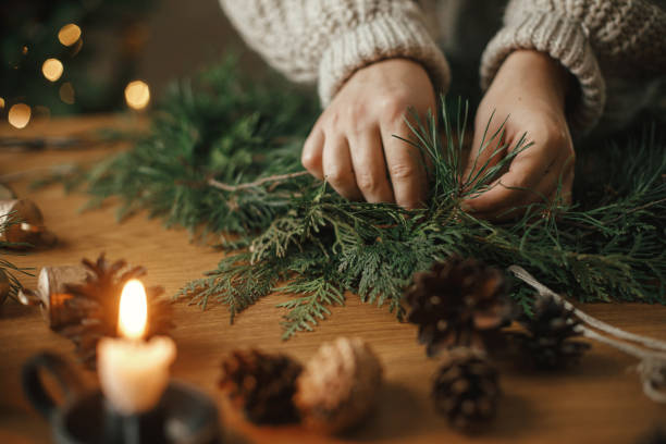faire une couronne rustique de noël en gros plan. mains tenant des branches de cèdre, faisant une couronne sur une table en bois avec des pommes de pin, une bougie, des cloches dans une salle de fête atmosphérique. préparatifs des vacances d’hiver - couronne de noel artisanale photos et images de collection