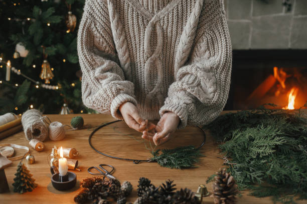 istock Making Christmas rustic wreath. Hands holding cedar branches, making wreath on wooden table with pine cones, candle, twine, bells in atmospheric festive room. Winter holiday preparations 1709204480