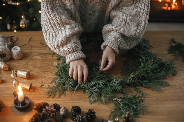 istock Making Christmas rustic wreath. Hands holding cedar branches, making wreath on wooden table with pine cones, candle, twine, bells in atmospheric festive room. Winter holiday preparations 1709205000
