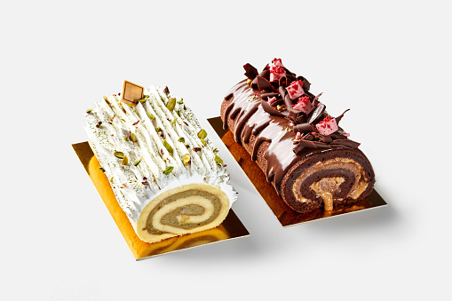Two Swiss cake rolls of vanilla and chocolate biscuit with pistachio custard, chocolate buttercream and caramel garnished with whipped cream, nuts, ganache and berry marshmallow on golden cardboard