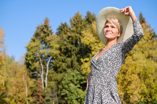 adult beautiful woman in a dress and hat against the background of autumn trees and blue sky on a sunny day
