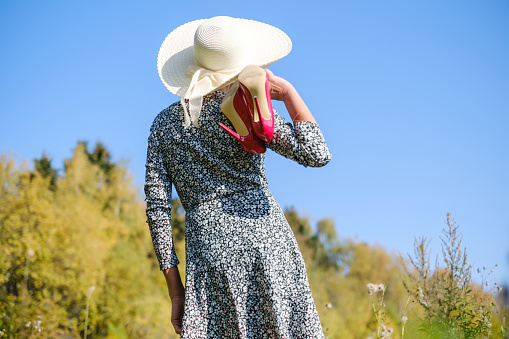adult beautiful woman with shoes in her hands in a dress and hat against the background of autumn trees and blue sky on a sunny day