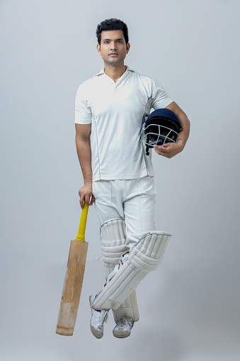 Full body portrait of Man in cricketer dress Holding helmet and camera in hand looking towards the camera