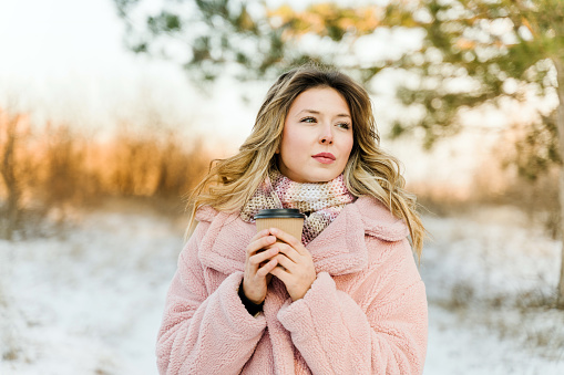 Beautiful woman drinking hot coffee in winter forest during sunset