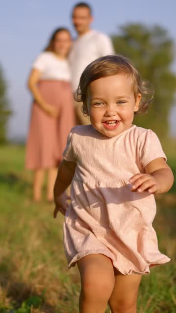 Expectant Couple Watching Cheerful Little Girl Walking on Footpath amidst Grassy Field on Sunny Day - VERTICAL