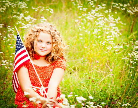 Beautiful 11 year old girl with an American flag sitting in a fairy tale field of wildflowers