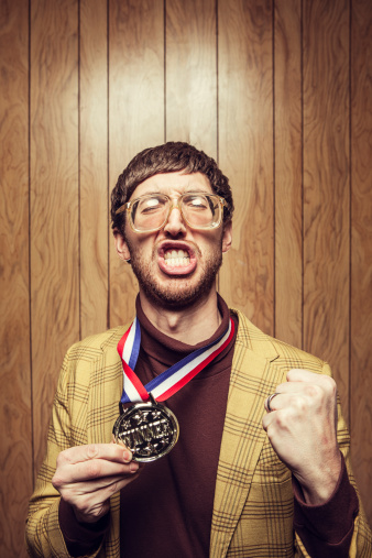 A nerd in a turtleneck and ugly yellow blazer with oversized glasses, bad hair, and a funny face celebrates proudly in front of retro wood paneling with his gold medal prize for winning.  Vertical with copy space.